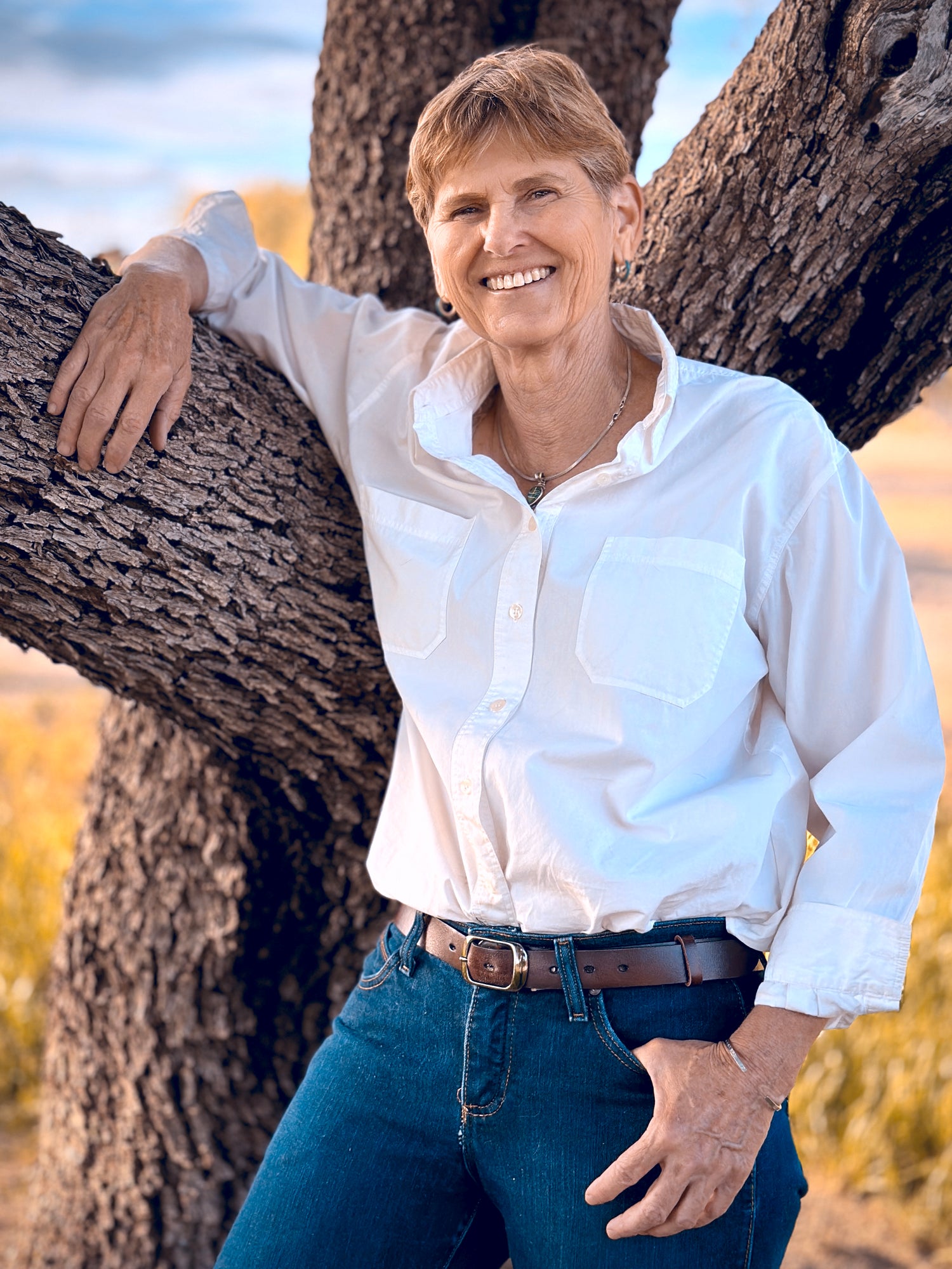 Alison, wearing a white shirt and jeans standing with her arm resting on a tree branch.