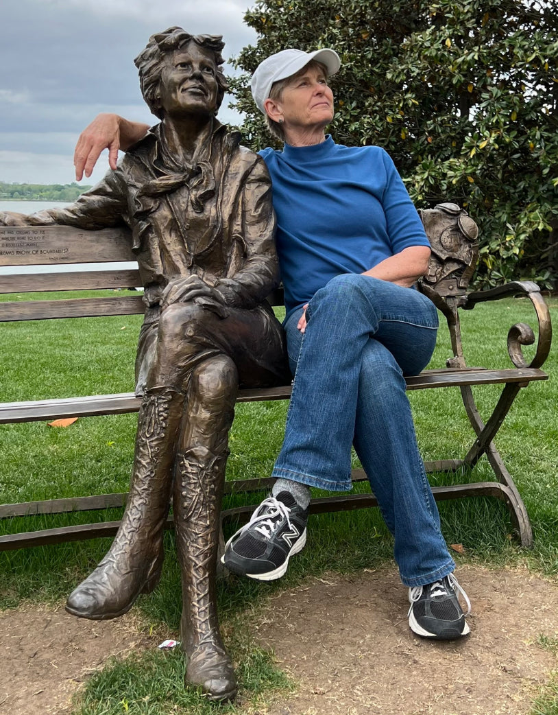 A photo of sapphic fiction author Alison Naomi Holt dressed in blue sitting on a  bronze bench with her arm around the shoulders of a bronze statue of strong heroine Amelia Earhart with them both looking into the sky